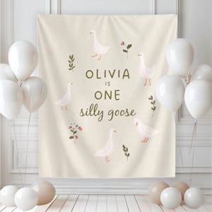May include: A beige backdrop with the text "OLIVIA IS ONE silly goose" and illustrations of white geese and floral accents. White and beige balloons are arranged around the backdrop, creating a festive atmosphere.