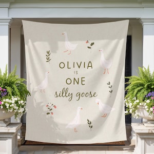 May include: A beige banner with the text "OLIVIA IS ONE silly goose" in a whimsical font. The banner is decorated with illustrations of white geese, sprigs, and small red flowers. The banner is hanging on a porch.