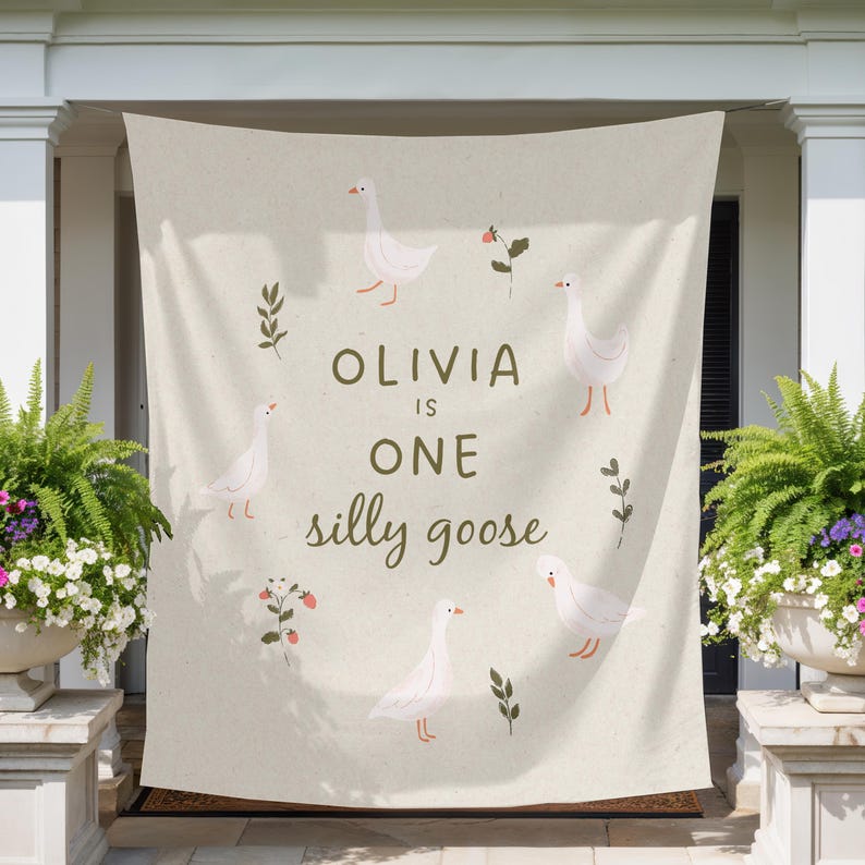 May include: A beige banner with the text "OLIVIA IS ONE silly goose" in a whimsical font. The banner is decorated with illustrations of white geese, sprigs, and small red flowers. The banner is hanging on a porch.