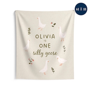 May include: A beige wall tapestry with the text "OLIVIA is ONE silly goose" in brown. The tapestry features illustrations of white geese, red berries, and green leaves. Dimensions are 68 x 80 inches.