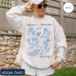 May include: White sweatshirt with a manatee graphic and the text "Advice from a Manatee." The design features blue manatees and aquatic plants with phrases like "breathe deep" and "live large."