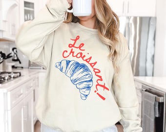 May include: A cream-colored sweatshirt with a blue and red graphic of a croissant and the text "Le Croissant!"