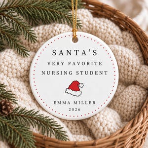 Może przedstawiać: Okrągła biała ceramiczna ozdoba z czerwoną kropkowaną obwódką i złotym sznurkiem do zawieszenia. Tekst brzmi SANTA'S VERY FAVORITE NURSING STUDENT, EMMA MILLER oraz 2026, z małą ilustracją czerwono-białej czapki Świętego Mikołaja.
