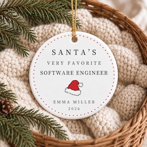 May include: A round white ceramic ornament with a gold hanging string. The text reads SANTA'S VERY FAVORITE SOFTWARE ENGINEER, EMMA MILLER, and 2026. A small red and white Santa hat graphic is centred between the text and the name.