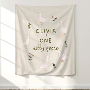 May include: A beige fabric wall hanging featuring a whimsical design. The design includes white geese, green sprigs, and red berries arranged around the text "OLIVIA IS ONE silly goose".