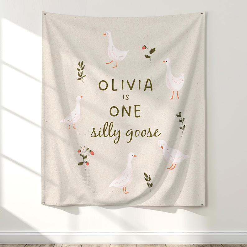 May include: A beige fabric wall hanging featuring a whimsical design. The design includes white geese, green sprigs, and red berries arranged around the text "OLIVIA IS ONE silly goose".