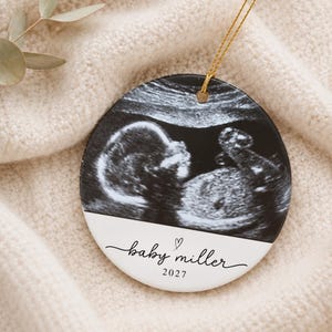 May include: A round ceramic ornament featuring a black and white ultrasound image of a fetus. The bottom section is white with the black text "baby miller" written in a cursive script, a small heart icon, and the year "2027" printed below.
