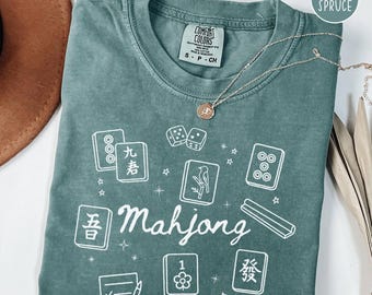 May include: Blue spruce Comfort Colors t-shirt with a white Mahjong graphic. The shirt features various Mahjong tiles, dice, and the word "Mahjong" in a script font. A silver necklace with a pendant is draped across the shirt.