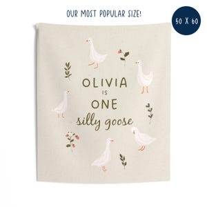 May include: A beige tapestry with the text "OLIVIA is ONE silly goose" surrounded by illustrations of white geese, leaves, and flowers. The tapestry is 50 x 60 inches and has the text "OUR MOST POPULAR SIZE!" at the top.