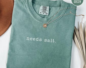 May include: A light green t-shirt with the phrase "needs salt" in white lettering. The shirt is folded and has a Comfort Colors tag. A silver necklace with a round pendant rests on the shirt. The words "ships fast!" are in a blue rectangle.