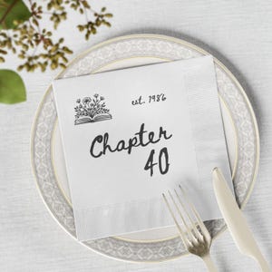 May include: A square white paper napkin featuring a black printed graphic of an open book with flowers emerging from the pages. The text reads "est. 1986" and "Chapter 40" in a black script font. The napkin is placed on a decorative plate with a fork and knife.