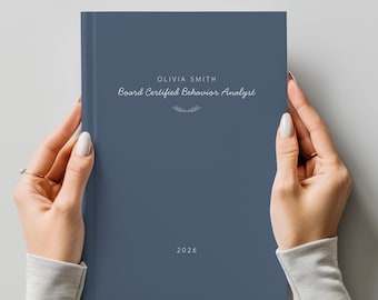 May include: A slate blue hardbound planner featuring the text OLIVIA SMITH and Board Certified Behavior Analyst in white, with a small decorative flourish below the text and the year 2026 printed in white at the bottom center of the cover.