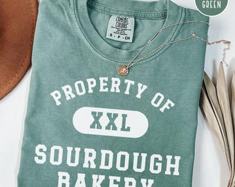 May include: A light green t-shirt with the text "PROPERTY OF XXL SOURDOUGH BAKERY" in white. The shirt has a crew neck and short sleeves. A silver necklace with a pendant is draped across the shirt.