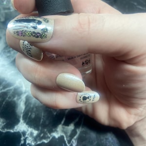 Pode incluir: Close-up de uma mão com esmalte de unha cor nude e um design de silhueta de mulher em cada unha. O frasco de esmalte de unha é visível no fundo com o texto "Made in USA" no rótulo.