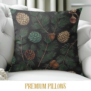 May include: A decorative pillow with a dark background featuring a pattern of pine needles, pine cones, and acorns in shades of green, brown, and tan. The pillow is square and rests on a white couch. The text "PREMIUM PILLOWS" is at the bottom.