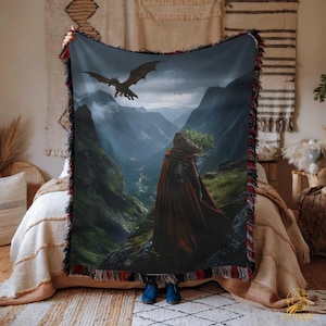 Puede incluir: Una manta tejida que presenta un paisaje de fantasía con un dragón volando sobre una figura con una capa roja. La manta tiene un fondo azul oscuro con una cordillera y un fleco rojo y negro.