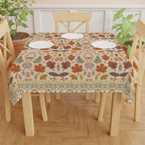 May include: A rectangular tablecloth with a beige background and a repeating pattern of autumn leaves in shades of orange, brown, and green. The border features an ornate design with blue, yellow, and green accents. Three white plates are on the table.