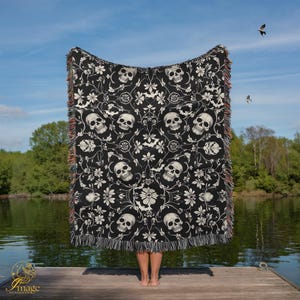 Puede incluir: Manta negra con un patrón repetido de calaveras blancas y diseños florales. La manta tiene un borde con flecos multicolores. La manta se sostiene frente a una persona de pie en un muelle de madera.