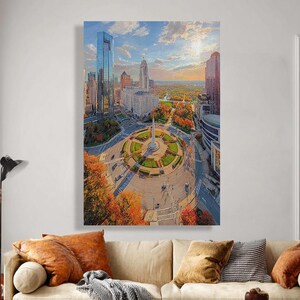 May include: A large vertical canvas print of a city skyline with a central monument and circular park. The image features tall buildings, a blue sky with clouds, and trees with autumn foliage. The artwork is displayed above a beige sofa.