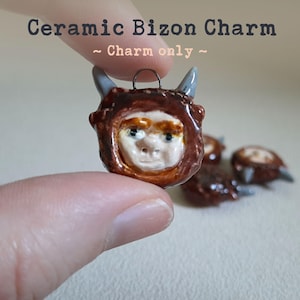 May include: A ceramic charm shaped like a brown furry creature with a human-like face. The charm has two small gray horns and is a handmade item.