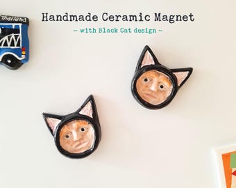 May include: Two handmade ceramic magnets with a black cat design. Each magnet features a black cat face with pink ears and a light brown human-like face.