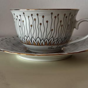 May include: A white teacup and saucer with a gold rim and black and gold floral design. The teacup has a black and gold floral design and the text "Je t'aime" written on the bottom.