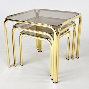 May include: A set of three nesting tables featuring polished gold-tone metal frames with rounded corners and square, smoked glass tops. The tables are stacked together, showcasing a tiered arrangement with reflective surfaces and metallic legs.