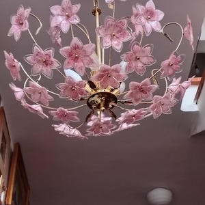 以下が含まれることがあります： 金色のフレームと、ピンク色の花型の照明器具が多数取り付けられた、花のシャンデリア。花は半透明で、濃いピンク色のアクセントがあります。白い天井に取り付けられています。
