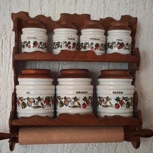 May include: A two-tiered wooden wall shelf holding seven ceramic canisters with brown lids and a vegetable pattern. The canisters are labeled with the text: VEGETA, KAKAO, PAPRIKA, KAVA, SO, BRAŠNO, and ŠEĆER. A wooden paper towel holder is attached below.