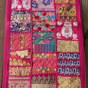 May include: A vibrant pink textile with a patchwork design. The rectangular piece features various embroidered squares in shades of pink, gold, and teal. The intricate patterns include floral and geometric motifs, creating a rich, textured appearance. The edges are framed with a solid pink border.