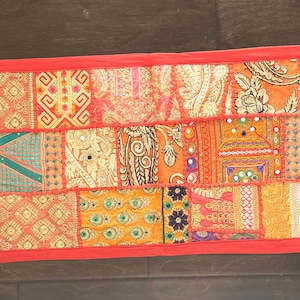 May include: A red table runner with a patchwork design of colorful fabric squares. The runner is decorated with gold embroidery and sequins. It has tassels on each end.