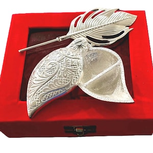 May include: A silver-colored metal leaf-shaped trinket box with an intricate embossed pattern on the lid, accompanied by a matching silver-colored metal feather-shaped accessory, both presented inside a square red velvet-lined display box.