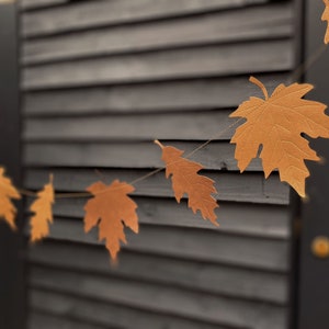 Könnte beinhalten: Girlande mit goldbraunen Ahornblatt-Ausschnitten. Die Blätter sind mit detaillierten Adern versehen und an einer Schnur aufgereiht. Der Hintergrund ist dunkel und geriffelt, was eine herbstliche Atmosphäre erzeugt.
