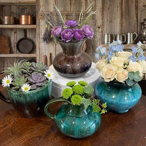 May include: A collection of four ceramic vessels featuring glazed finishes in shades of deep brown, teal, and green. The vessels contain arrangements of purple ranunculus, cream roses with blue delphiniums, green chrysanthemums, and assorted succulents.