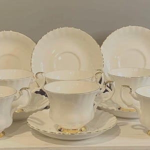 May include: A six-piece tea set featuring white teacups and saucers with gold detailing. The cups have decorative handles and sit on matching saucers. The edges of the plates and cups are scalloped. The teacups have a gold base.