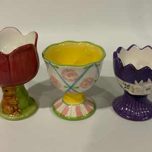May include: Three decorative ceramic egg cups. One is red and shaped like a tulip with a bunny base. The second is white with pink roses and blue stripes. The third is purple with bunny and floral details.