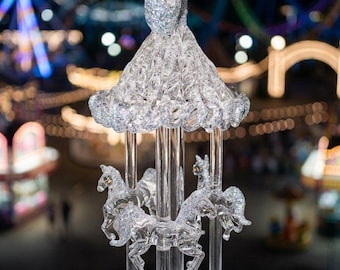 May include: A clear acrylic carousel ornament featuring multiple horses with silver glitter accents. The structure includes a central pole, a decorative canopy top, and a circular base, all crafted from transparent material with sparkling details.