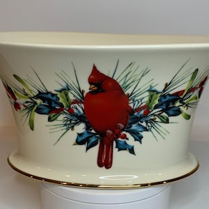 May include: Cream ceramic bowl with a gold rim, featuring a red cardinal bird illustration surrounded by holly and pine branches. The bowl has a flared shape and a decorative base.