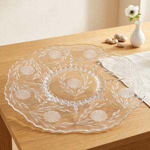 May include: A clear glass serving platter featuring a scalloped edge and a central circular section. The surface is decorated with etched floral motifs, including a central flower surrounded by six smaller flowers and leafy stems.