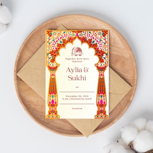Puede incluir: Una tarjeta de invitación con un diseño ornamentado, un motivo de elefante y los nombres "Aylia & Sukhi". La tarjeta incluye la fecha "04 de noviembre de 2024" y la hora "9 am | seguido del almuerzo". Se encuentra sobre una bandeja de madera con un sobre marrón.