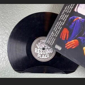 May include: A black vinyl record resting in a curved holder. The record's label reads "The Vinyl Resting Place." A record album cover is partially visible, displaying song titles and a parental advisory warning.