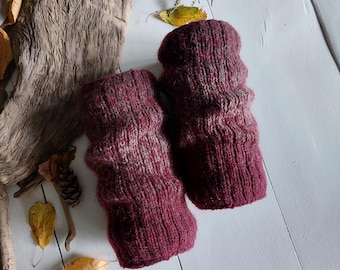 May include: A pair of burgundy-colored knitted wrist warmers. The warmers are made of a textured yarn with a ribbed design. The background includes dried leaves, wood, and sprigs of flowers, suggesting a fall theme.