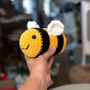 May include: A crocheted bee plush toy with yellow and black stripes, white wings, and black eyes. The bee is being held in a hand.