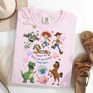 May include: A light pink t-shirt featuring cartoon characters from the movie Toy Story. The shirt has Buzz Lightyear, Woody, Jessie, Rex, Slinky Dog, Hamm, Bullseye, and an alien. The text "you've got a friend in me" is also on the shirt.