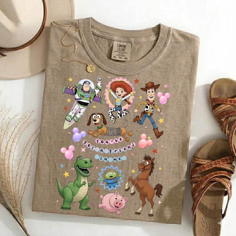 May include: A tan t-shirt featuring colorful cartoon characters from the Toy Story movie. The shirt includes Buzz Lightyear, Jessie, Woody, Rex, Slinky Dog, and other characters. The text "you've got a friend in me" is also on the shirt.