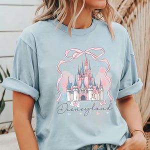 Op de afbeelding: Lichtblauw T-shirt met een roze en blauwe illustratie van een kasteel en het woord "Disneyland" in script. Het kasteel is omlijst door een roze lint. De mouwen zijn opgerold.