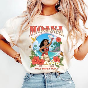 May include: Cream-colored t-shirt featuring a colorful graphic of Moana from Walt Disney World. The design includes the word "MOANA" in a retro font, a tropical scene, and the text "EST 2016". The shirt is adorned with floral and butterfly accents.