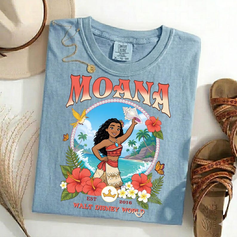 May include: Light blue t-shirt featuring the word "MOANA" in a retro font. The shirt has a graphic of Moana holding a seashell, surrounded by flowers and butterflies. The text "EST 2016 WALT DISNEY WORLD" is also visible.