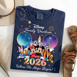 May include: Navy blue t-shirt featuring Disney characters Mickey, Minnie, Donald, Daisy, Goofy, and Pluto. The shirt has the text "Disney Family Vacation 2026 Where The Magic Begins!" and is surrounded by fireworks and stars.