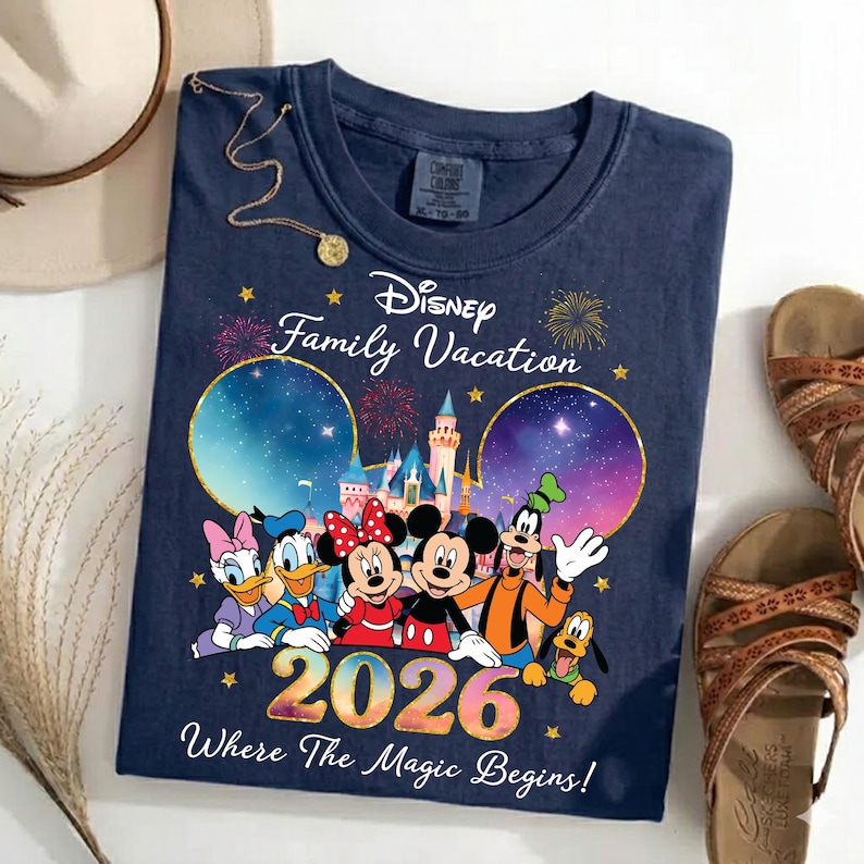 May include: Navy blue t-shirt featuring Disney characters Mickey, Minnie, Donald, Daisy, Goofy, and Pluto. The shirt has the text "Disney Family Vacation 2026 Where The Magic Begins!" and is surrounded by fireworks and stars.